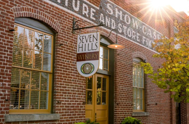 A brick building with arched windows and a sign reading “Seven Hills Winery”; sunlight shines above, and the building is partially covered by a yellow-leaved tree and painted advertisements.