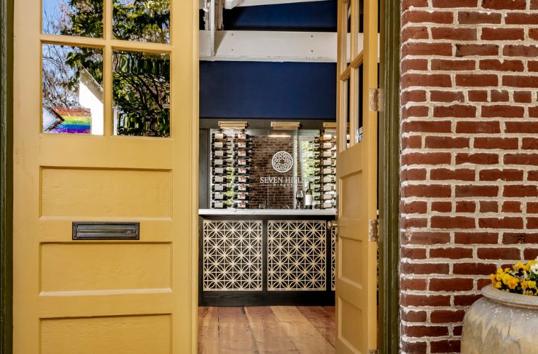 Open yellow double doors reveal a stylish interior with a wall-mounted wine rack, blue accents, decorative paneling, and a brick wall on the right. A barrel planter with yellow flowers is partially visible in the foreground.