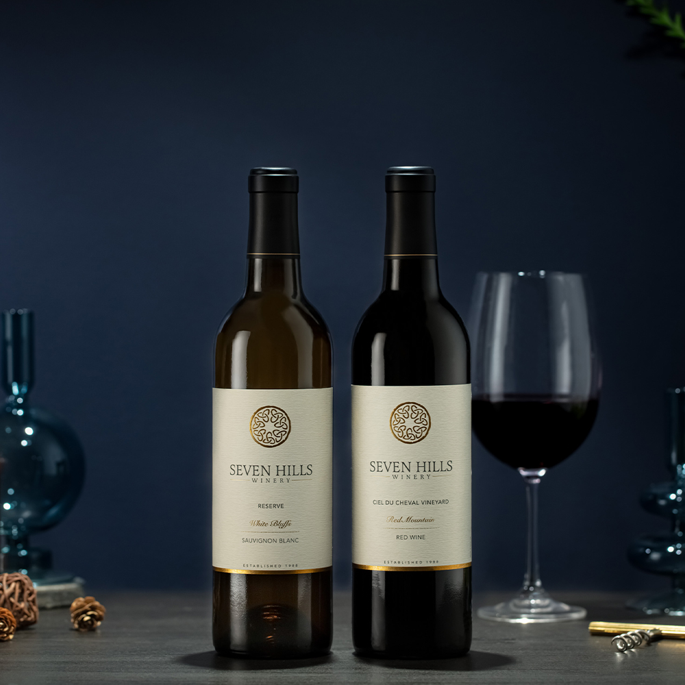 Two bottles of Washington's Finest Wine from Seven Hills—Sauvignon Blanc and Red—stand side by side on a table. A wine glass filled with red wine and blue vases create an elegant backdrop, making this the perfect wine gift.
