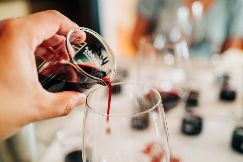 A hand pours red wine from a small glass pitcher into a wine glass, with other wine glasses and a blurred background visible.