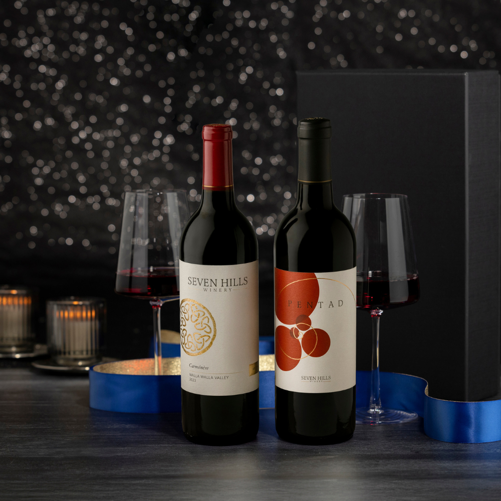 Two bottles of Washington's Finest Wine with glasses, a blue ribbon, candles, and a black gift box create the perfect wine gift on a table set against a festive, glittering dark background.