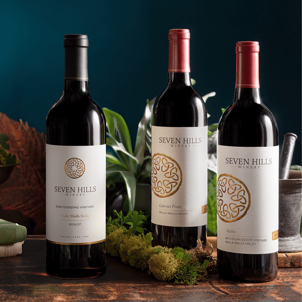 Three bottles of the 2022-bordeaux-trio from Seven Hills Winery stand on a table with greenery and moss, set against a dark blue background. Each bottle features a white label with gold accents and the Seven Hills logo.