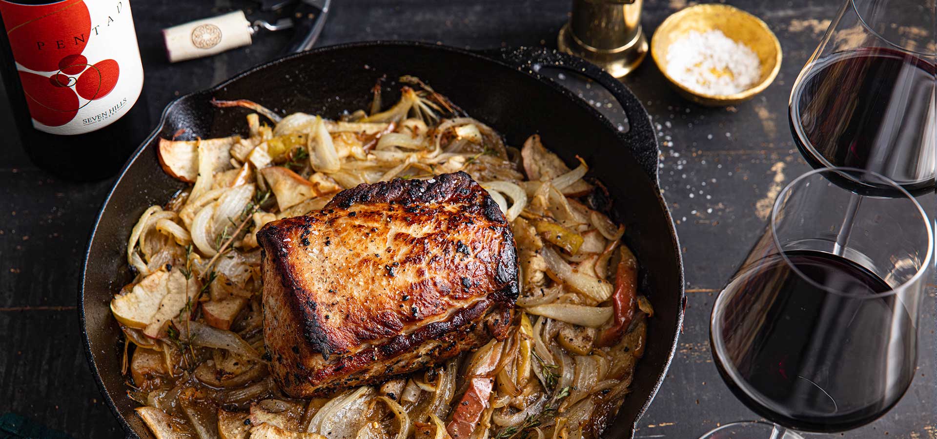 A roasted pork loin sits on a bed of caramelized onions and apples in a cast iron skillet, next to two glasses of red wine, a bottle of wine, a gold bowl of salt, and a pepper grinder on a dark table.