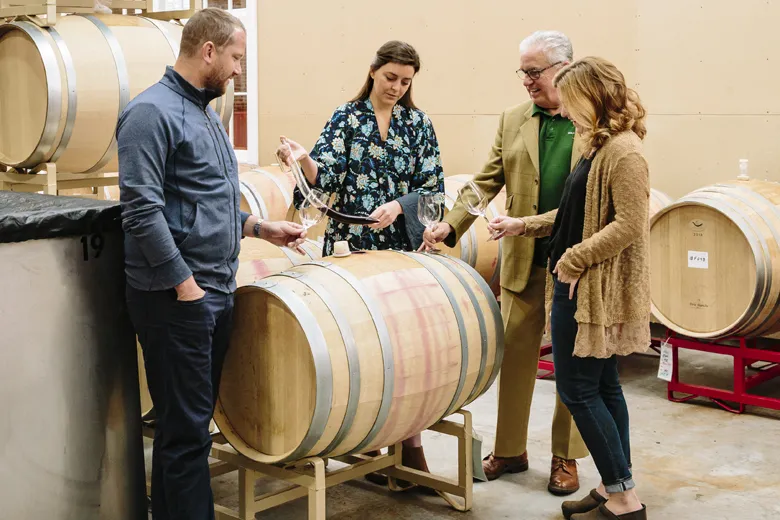 Four people stand around a wine barrel in a winery, holding wine glasses and sampling wine. Two men and two women are engaged in conversation, surrounded by several large wooden barrels.