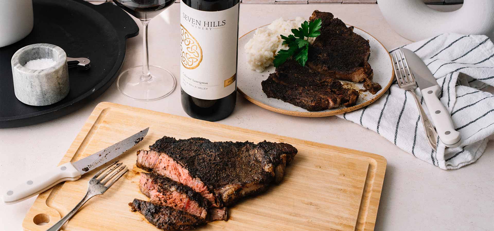 Sliced, medium-rare steak on a wooden cutting board with a knife and fork. A plate of steak with mashed potatoes, a wine glass, and a bottle of red wine are in the background. A striped napkin sits nearby.