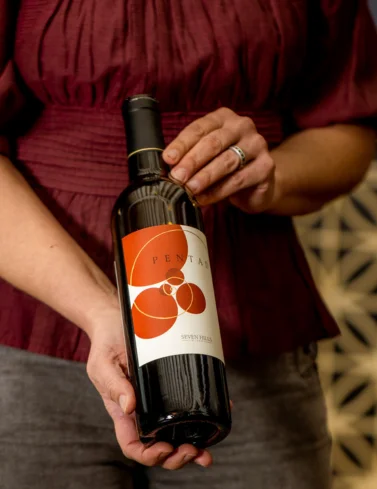 A person wearing a burgundy top and gray pants holds a bottle of wine with a modern, orange geometric label that reads Penta and Seven Peaks.