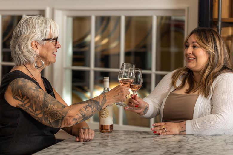 Two women sit at a marble table, smiling and clinking glasses of rosé wine. One woman has grey hair, tattoos, and glasses; the other has long brown hair and wears a white cardigan. A wine bottle is on the table.