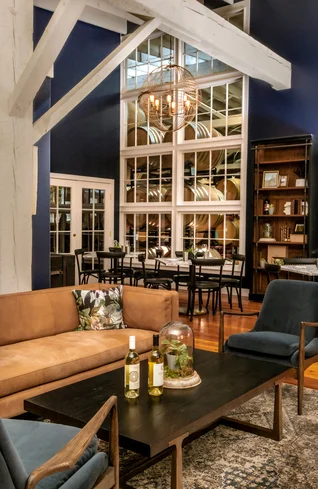 Modern lounge with a tan sofa, blue chairs, and a black coffee table holding wine bottles. Large windows reveal wine barrels in another room. A chandelier hangs above a dining area with a dark blue accent wall.