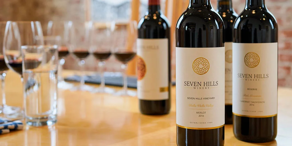 Four bottles of Seven Hills Winery red wine are displayed on a wooden table, accompanied by filled wine glasses. The background shows a softly lit, blurred indoor setting with a window.