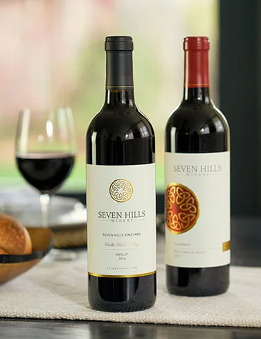 Two bottles of Seven Hills red wine stand on a table with a glass of red wine and a bread roll in the background. One label reads “Seven Hills Winery, Seven Hills Vineyard, Merlot 2018.”.