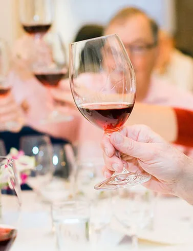A close-up of a hand holding a glass of red wine, raised in a toast at a table set with glasses and water, with other people blurred in the background doing the same.