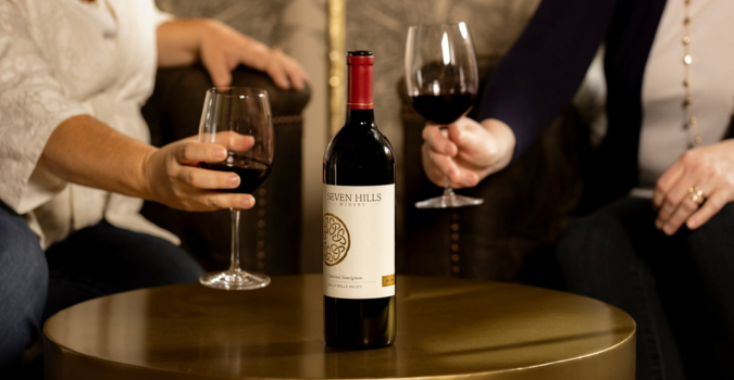 Two people seated on a couch hold glasses of red wine, with a bottle of Seven Hills wine placed on a round, gold-colored table between them.