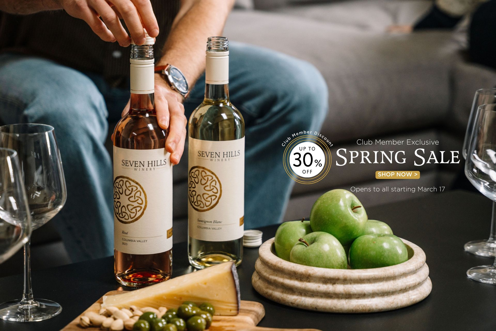 A person sets two bottles of Seven Hills wine on a table next to green apples, cheese, olives, nuts, and empty wine glasses. Text advertises a spring sale with up to 30% off, starting March 17.