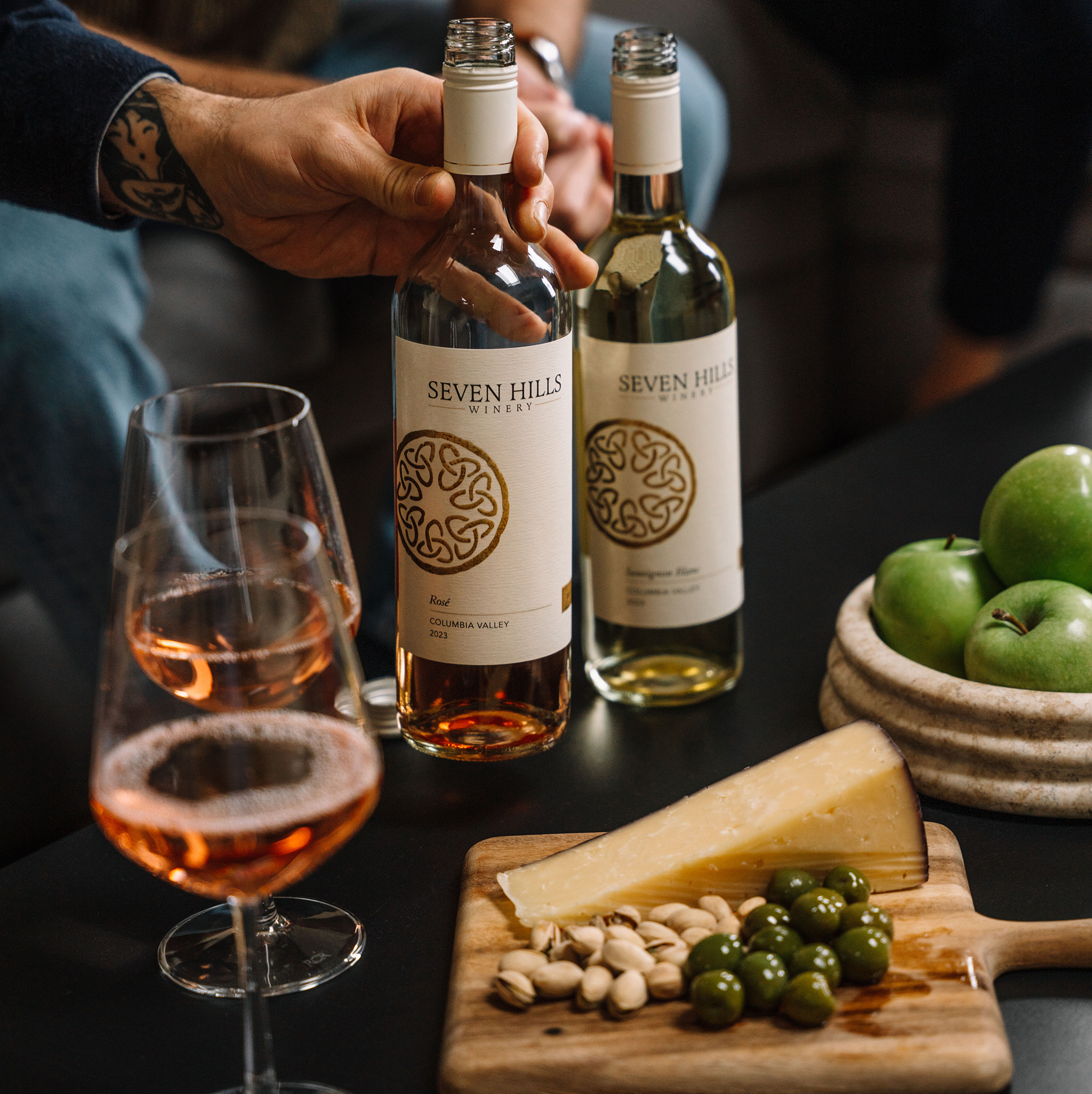 A hand holding a bottle of rosé wine labeled Seven Hills Winery next to another bottle of white wine, a glass of rosé, green apples, and a cheese board with cheese, olives, and nuts on a table.