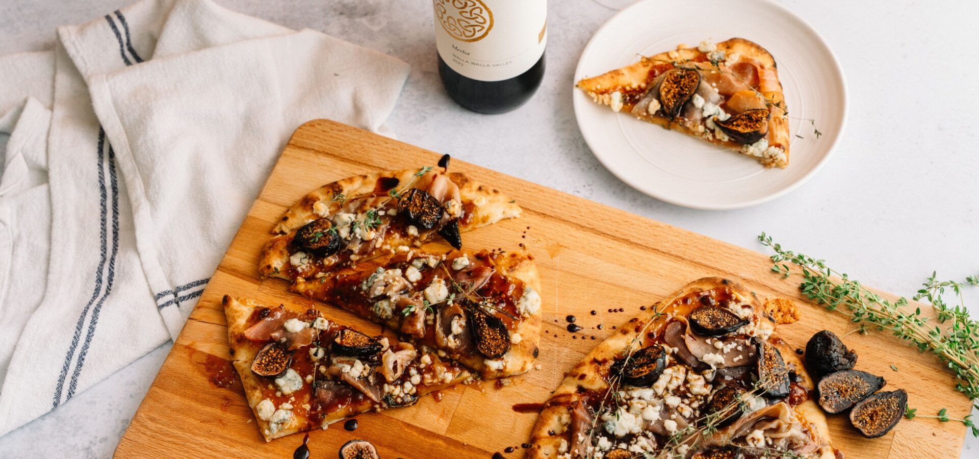 A wooden board with sliced flatbread topped with figs, cheese, and herbs. A plate with one slice and a bottle of wine are nearby, along with a white napkin and sprigs of fresh thyme on a light surface.