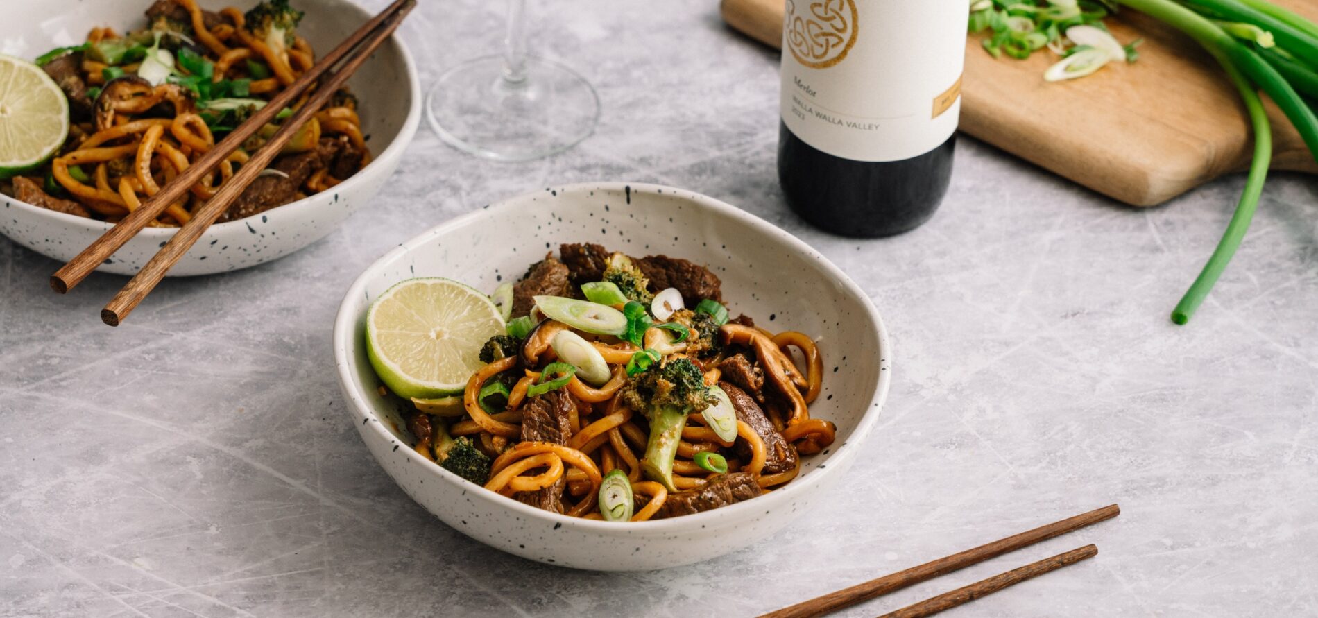 A bowl of stir-fried noodles with vegetables, beef, sliced scallions, and a lime wedge. Chopsticks rest beside the bowl, with another bowl, a wine bottle, and a cutting board with scallions in the background.