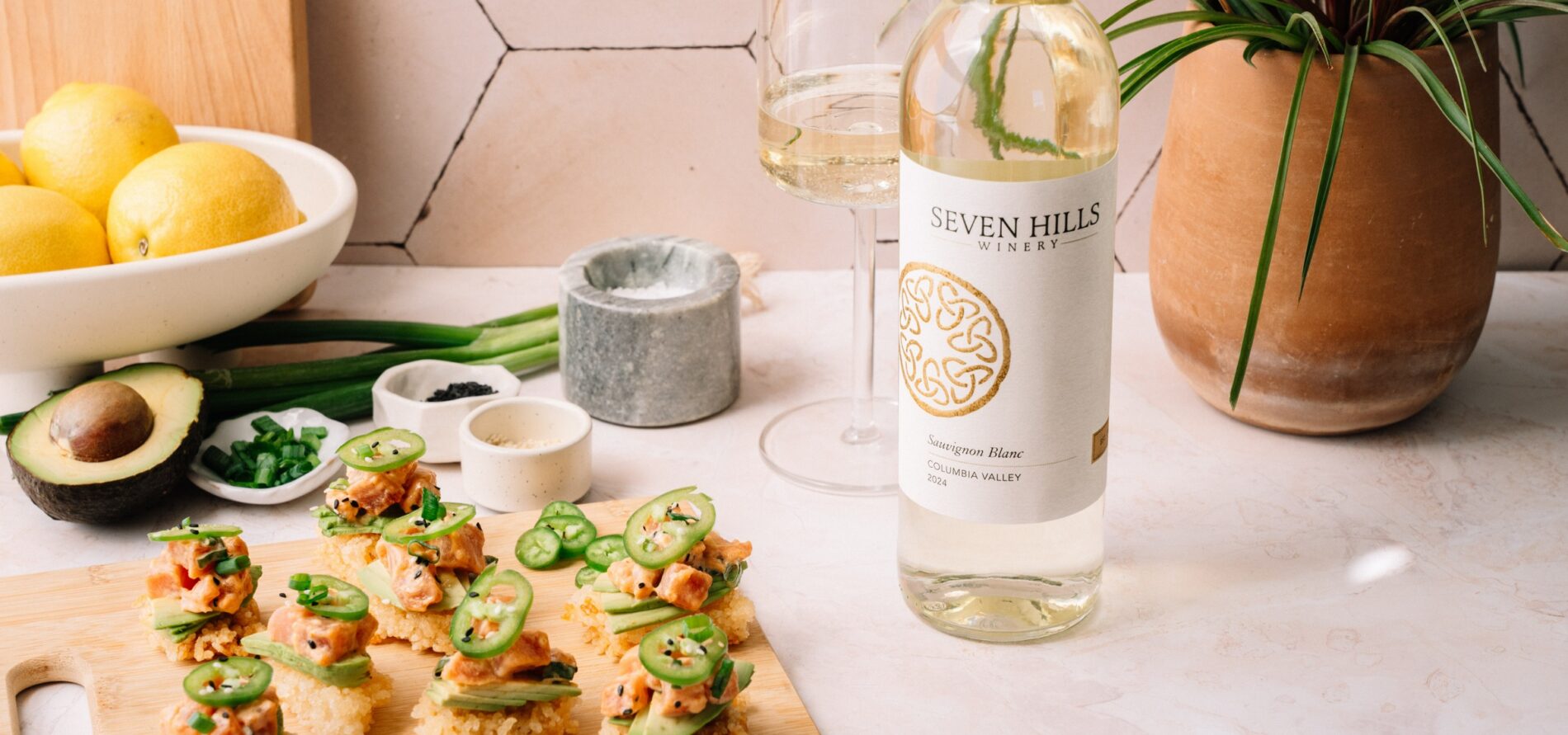 A bottle of Seven Hills white wine on a countertop next to a glass of wine, a potted plant, a bowl of lemons, sliced avocado, chopped green onions, and a wooden board with appetizers topped with jalapeños.