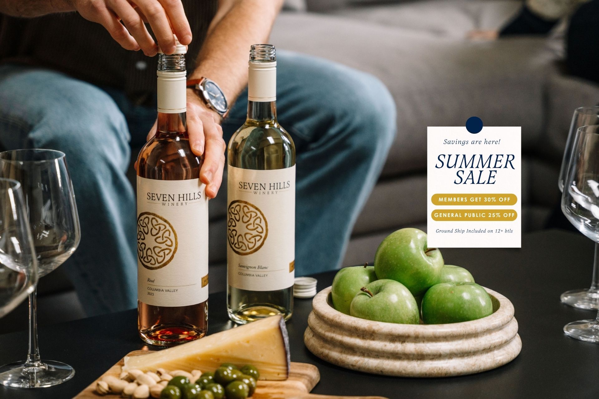 Two hands hold bottles of Seven Hills wine beside a plate of green apples, cheese, and olives. A sign advertises a summer sale: members get 25% off, general public 20% off, with ground shipping included.