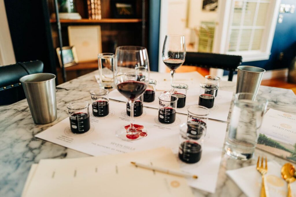 A marble table set for a wine tasting, featuring wine glasses, beakers with red wine, tasting sheets, and drinking water in metal and glass cups. Shelves and framed art are visible in the background.