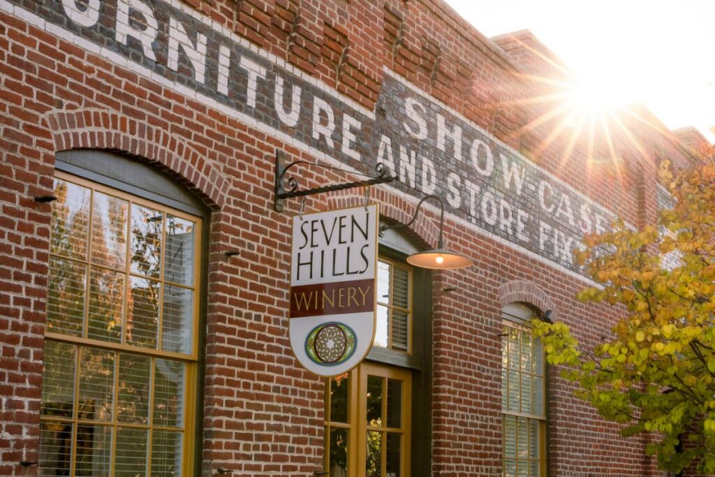 A brick building with old painted signs houses Seven Hills Winery. Sunlight shines through trees, reflecting off large windows.