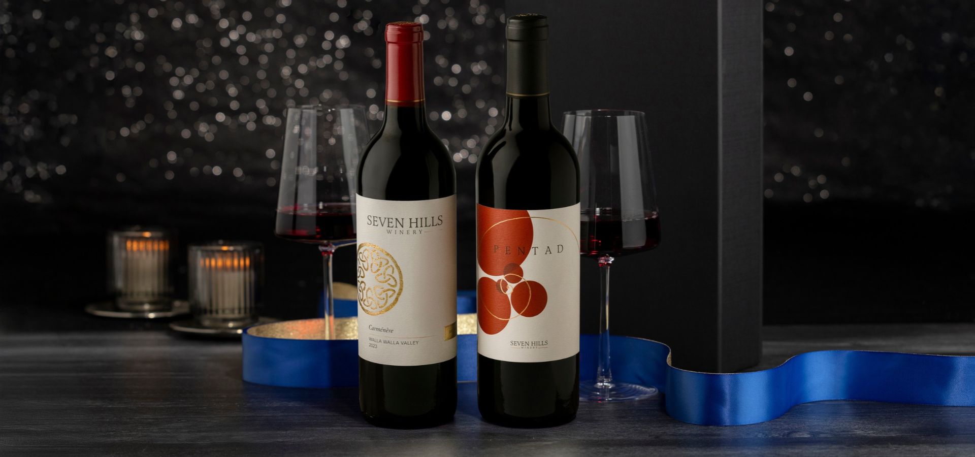 Two bottles of red wine with elegant labels stand beside filled wine glasses, candles, and a blue ribbon on a festive, dark, and sparkling background.